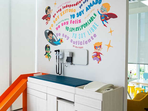Área de exploración con mesa pediátrica y un mural de superhéroes con frases de afirmación.
