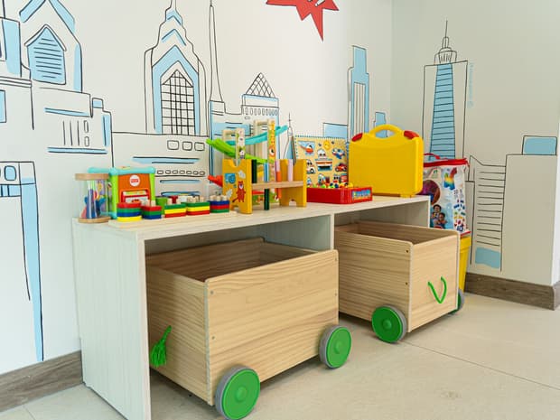 Zona de juegos con carritos de madera llenos de juguetes y un mural de ciudad.